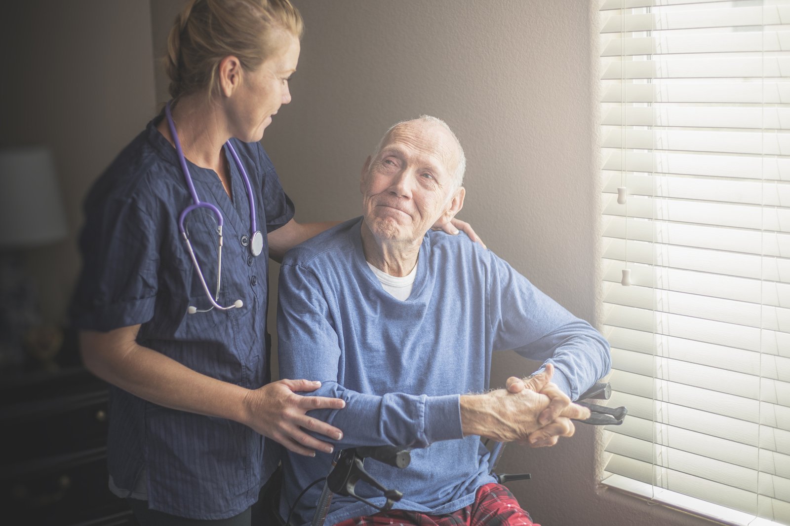 Ein kranker Mann sitzt am Fenster, während eine Pflegerin bei ihm steht. 