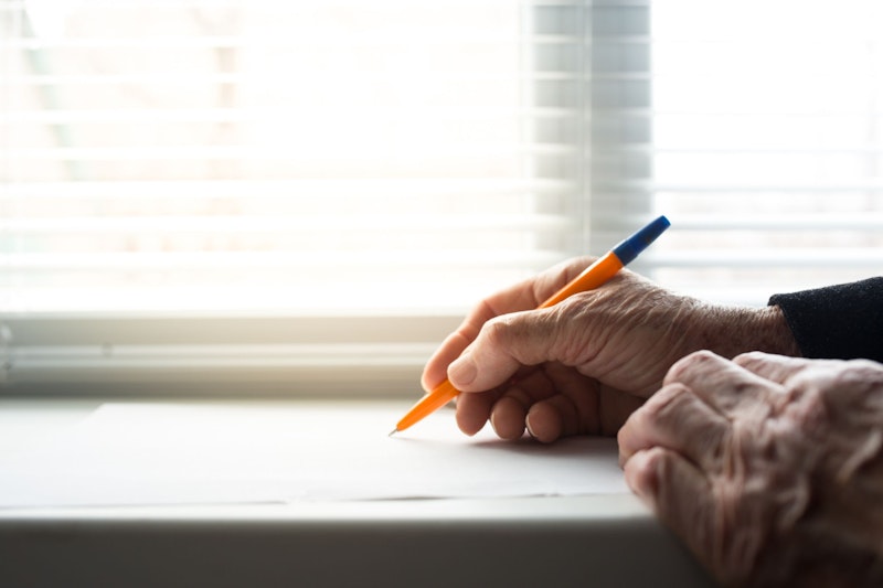 Das Bild zeigt die Hände eines Seniors, wie er ein Brief schreibt. 